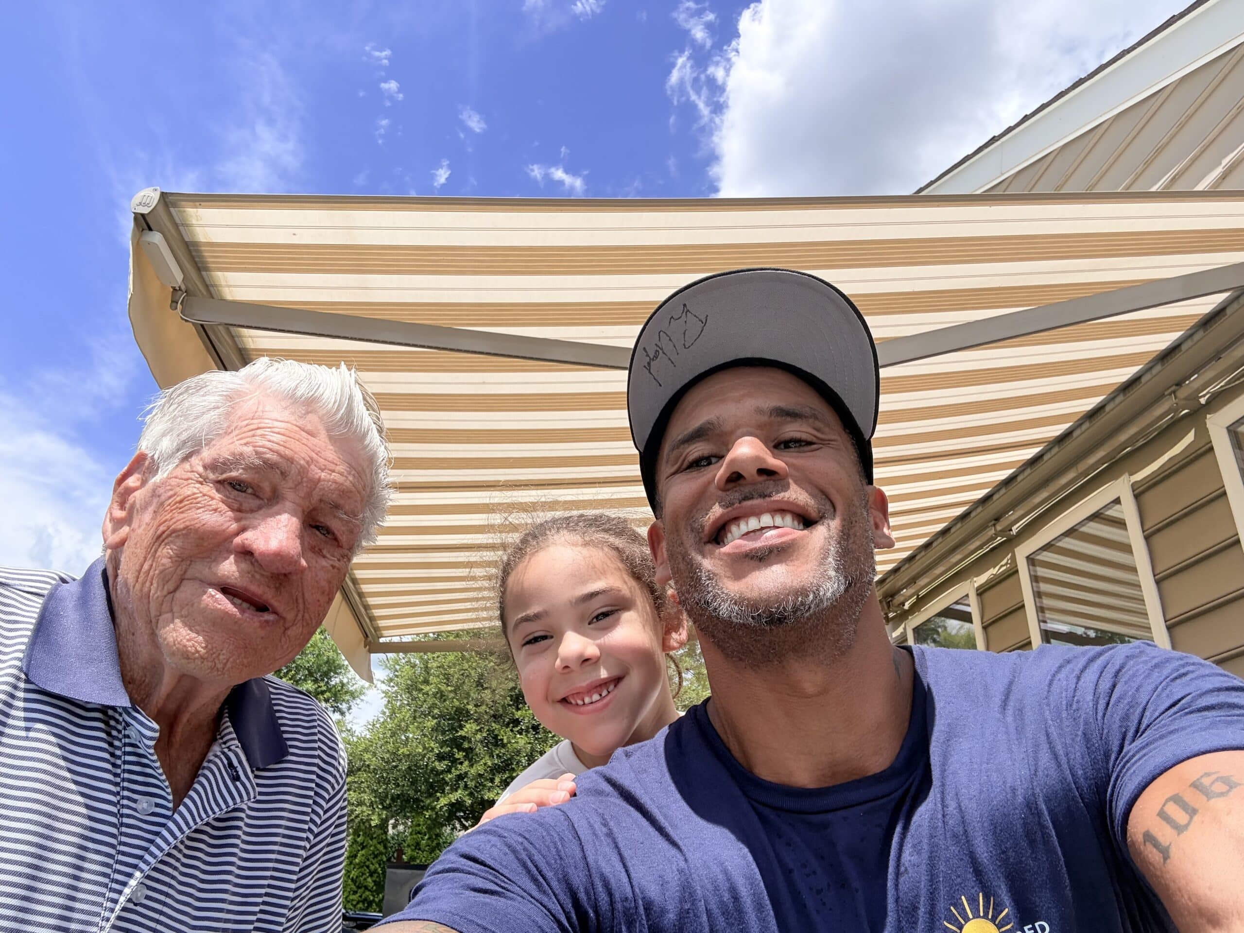 Three people—an older man, a young girl, and a man in a cap—smile outdoors under a striped awning on a sunny day. Green trees and part of a house are visible in the background.