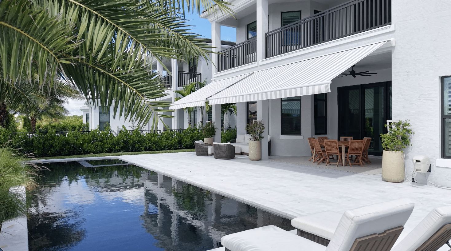 Modern white house with large windows, striped awning, patio furniture, and a swimming pool surrounded by palm trees and greenery under a blue sky.
